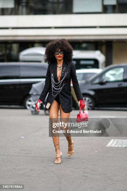 Guest wears black sunglasses, a silver chain with green stone pendant necklace, a black long blazer jacket with large chain pendant , a black V-neck...