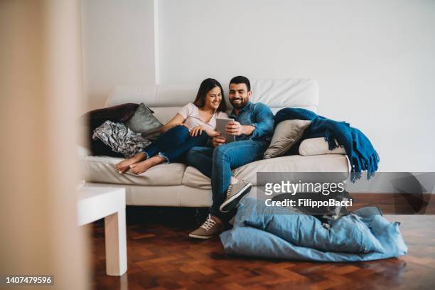 young adult couple together at home. they are sitting on the sofa using a tablet together - couple with ipad in home stock pictures, royalty-free photos & images