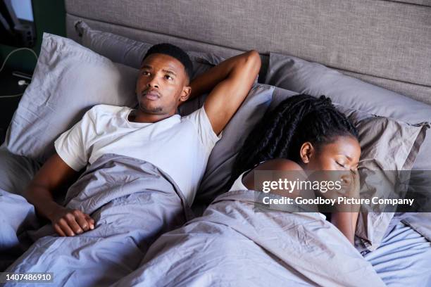 young man looking unhappy while lying in bed with his sleeping wife - husband stockfoto's en -beelden