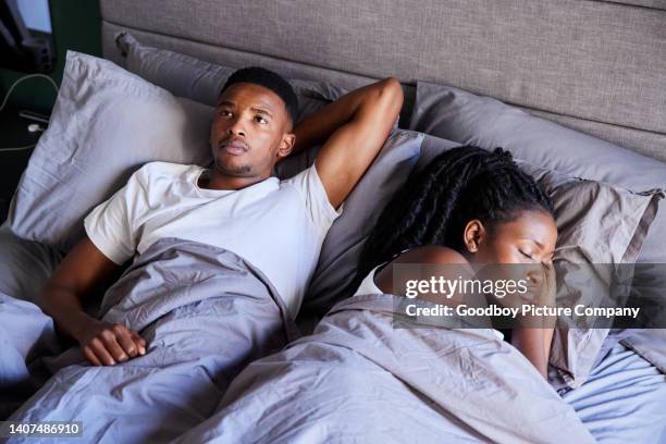 young man looking unhappy while lying in bed with his sleeping wife - couples in bed stock pictures, royalty-free photos & images