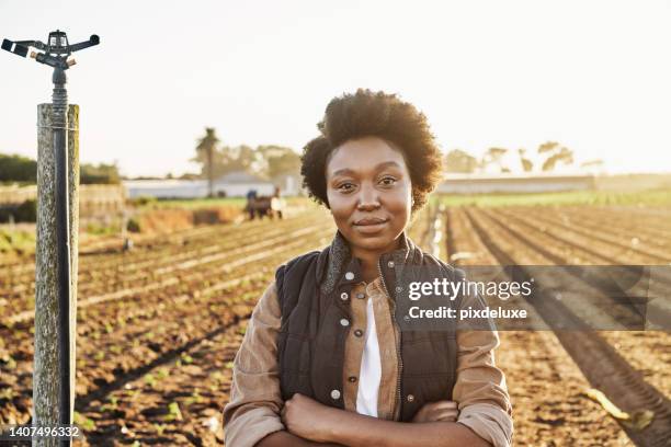 a young african female farmer in a coat on the farm on a sunny day. hispanic labor standing in a field with hands crossed in casual clothing. a black lady posing on a farm looking at a camera. - woman hand crossed stockfoto's en -beelden