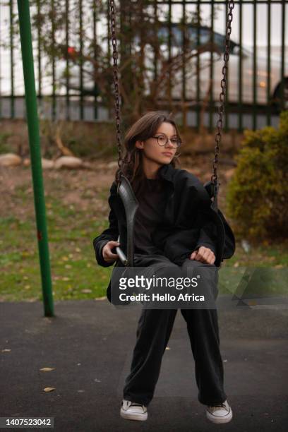 a beautiful young woman, a teenage girl with long flowing loosely worn brown hair and wearing lovely spectacles sits with an aloof or distracted expression in a swing seat in a green public park as she looks into the near distance - skimpy girls stock pictures, royalty-free photos & images