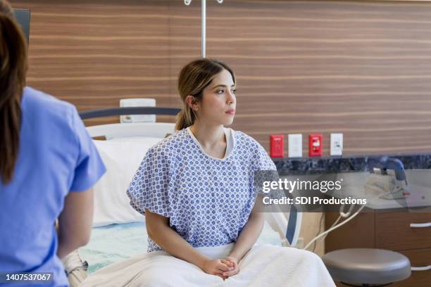 joven triste con bata de hospital se sienta en la cama - hospital gown fotografías e imágenes de stock