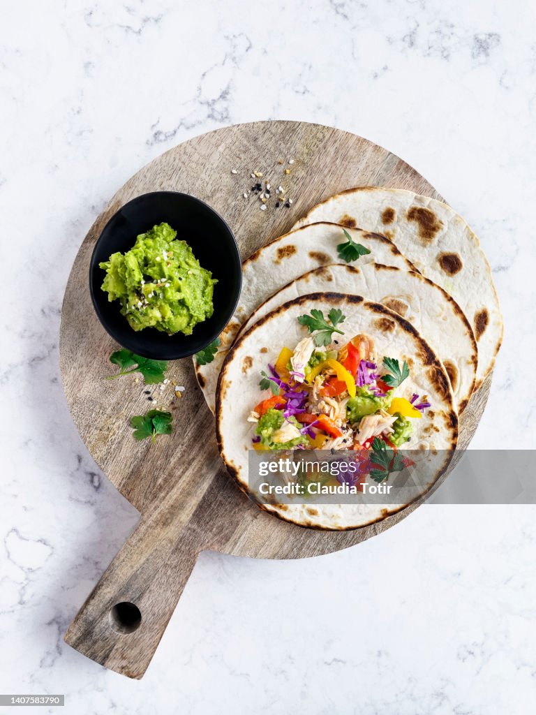 Chicken tacos, and a bowl of guacamole on wooden cutting board on white background