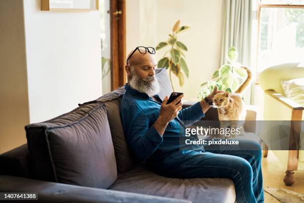 senior man using a phone to browse online while petting his cat on the couch at home. mature hipster male relaxing with his pet and scrolling on social media online on sofa. retired guy typing a text - cat hipster no stock pictures, royalty-free photos & images