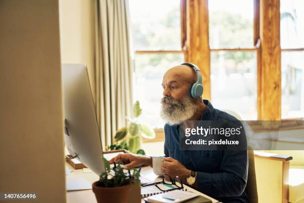 älterer mann, der kopfhörer trägt, um musik zu hören, während er zu hause an einem desktop-computer arbeitet. pensionierter mann, der online im internet surft und lieblingslieder genießt. reifer hipster-typ, der sich videos ansieht - mann anhimmeln stock-fotos und bilder