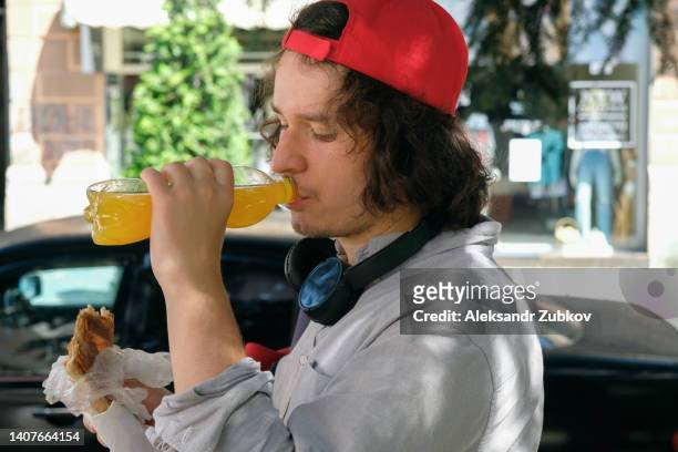 a man holds a georgian khachapuri in his hands, eats it, drinks carbonated water or a drink, outdoors. a student or tourist eats takeaway food, snacks. a teenager eats a puff pastry. the concept of fast food. getting to know georgian cuisine on a trip. - georgian man stock pictures, royalty-free photos & images