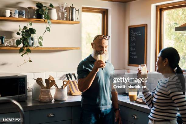 senior couple drinking healthy smoothies together in the kitchen at home. retired interracial husband and wife tasting a shake they made in the morning. mature man and woman enjoying a fresh snack - malt stockfoto's en -beelden