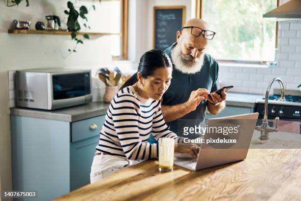 um casal maduro usando um laptop e telefone na cozinha em casa. marido e mulher navegando online usando aplicativos de mídia social com seus dispositivos sem fio em casa. homem e mulher navegando, comprando online - mixed media - fotografias e filmes do acervo