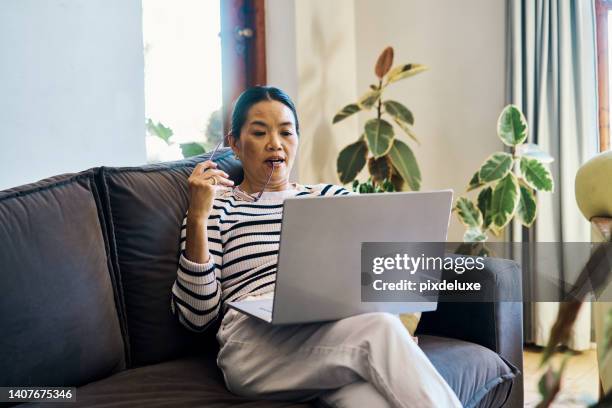 senior chinese woman reading an email on a laptop while sitting on the couch at home. one retired asian lady browsing the internet and streaming videos on the sofa. serious female searching online - one woman only videos stock pictures, royalty-free photos & images