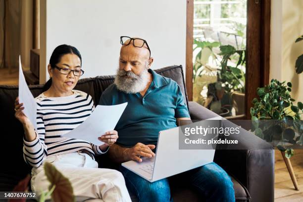 senior interracial couple using a laptop and going retirement documents online on the couch at home. mature husband and wife talking and reading paperwork while using a pc on the sofa in the lounge - pension 個照片及圖片檔