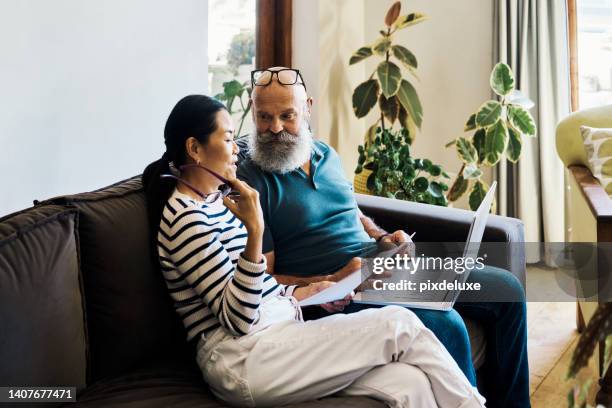 senior interracial couple using a laptop and going through paperwork to pay bills online on the couch at home. mature husband and wife talking and planning their retirement on a pc on the sofa - choosing insurance stock pictures, royalty-free photos & images