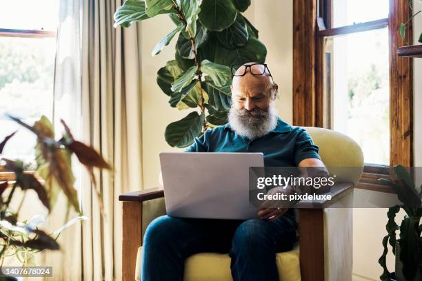 mature hipster male relaxing and scrolling on social media on a pc. retired guy watching series. senior man working on a laptop and searching the internet online while sitting on a chair at home. - man choosing stock pictures, royalty-free photos & images