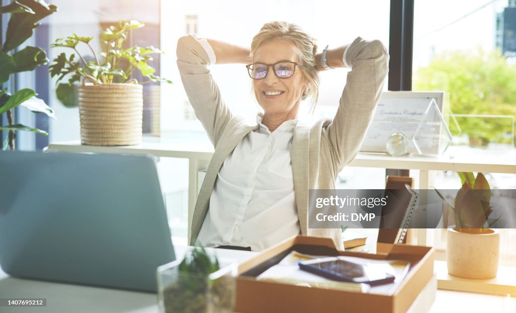 Happy mature manager satisfied and relieved to be done with deadlines and tasks. Business woman feeling accomplished and enjoying a relaxing break to stretch with hands behind her head in an office.