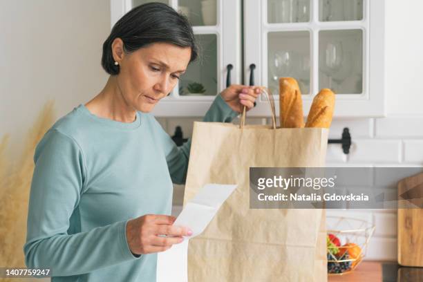 senior adult female worried while looking at receipt from grocery store. rise in price of products concept. buying consumer goods during inflation. facing economic downturn. kitchen unit and paper bag on background, horizontal. closeup - receipt 個照片及圖片檔