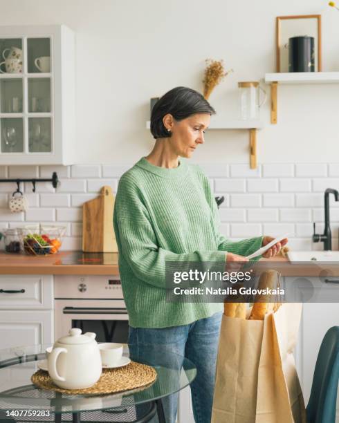 concerned senior female looking at receipt from supermarket. shocking rise in price of products concept. buying consumer goods during crisis and inflation. facing economic downturn. shopping paper bag on background, vertical - pay rise stock-fotos und bilder