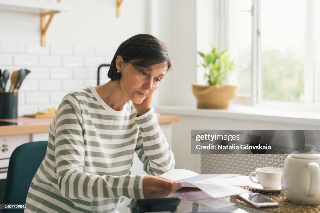 Upset woman thinking about high prices while looking at utilities, gas, electricity, rental charges, water bill due to inflation and crisis. Planning personal budget while sitting in kitchen. Weighing options on how to save money