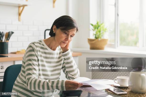 upset woman thinking about high prices while looking at utilities, gas, electricity, rental charges, water bill due to inflation and crisis. planning personal budget while sitting in kitchen. weighing options on how to save money - rechnungen stock-fotos und bilder