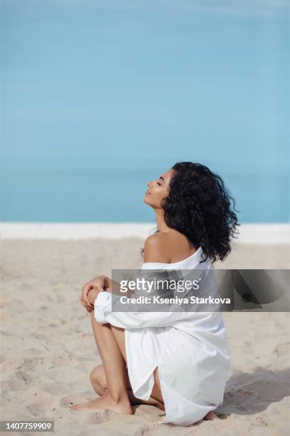 young beautiful mixed race woman with long curly hair sits in a white shirt on a sandy beach, breathes in the sea breeze and smiles - beautiful armenian women fotografías e imágenes de stock