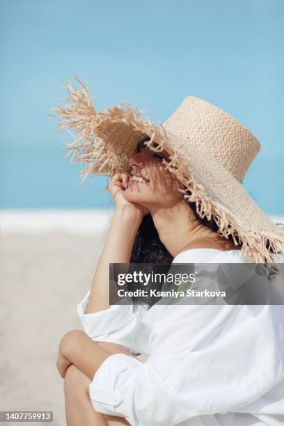 young beautiful mixed race woman with long curly hair sits on the beach in a straw hat with her eyes closed, looks at the sun and smiles - chapeau de paille photos et images de collection