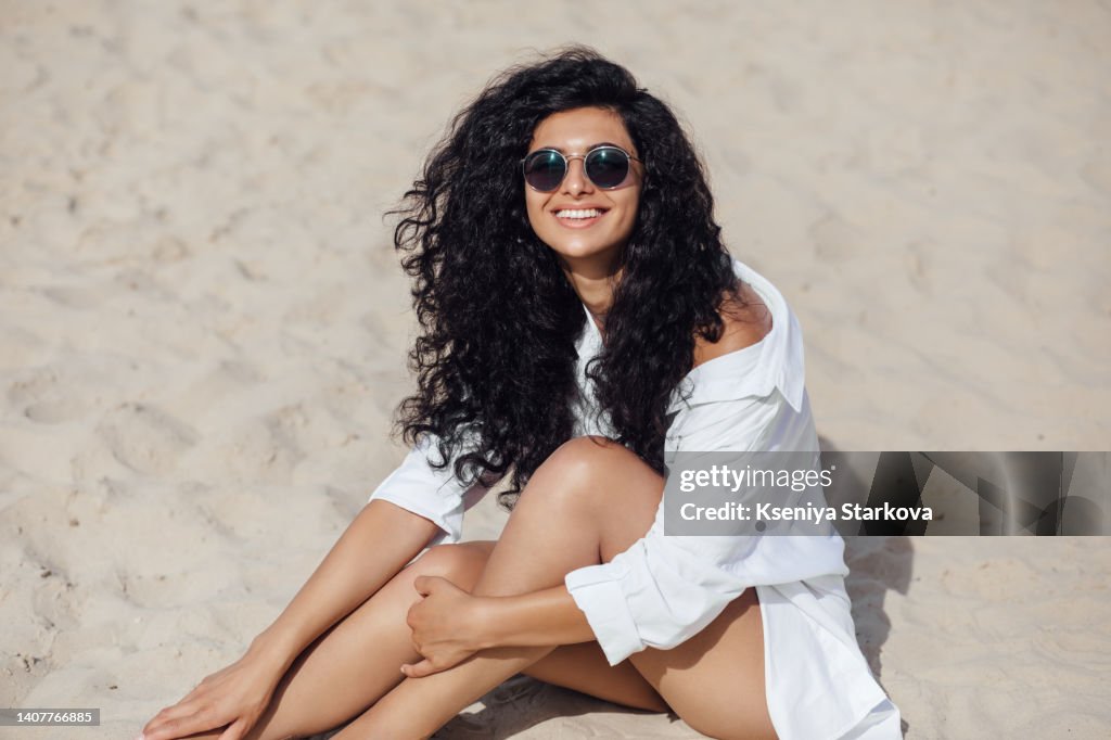 Young Beautiful Mixed Race Woman With Long Curly Hair Sits On A Sandy Beach  In A White Shirt And Sunglasses Looks At The Camera And Smiles High-Res  Stock Photo - Getty Images
