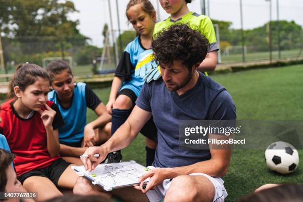 niños de un equipo de fútbol escuchando a su entrenador, sentados juntos en un campo de fútbol - entrenador fotografías e imágenes de stock
