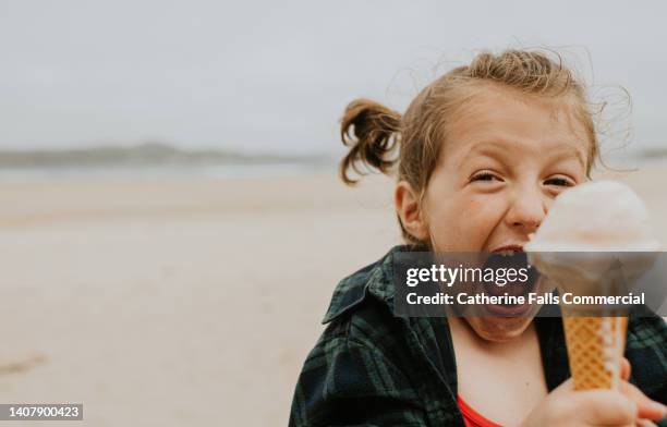 ecstatic little girl holds a vanilla ice-cream on a beach, looking delighted as she holds the cone towards the camera. - girl eating messy ice cream cone stock-fotos und bilder