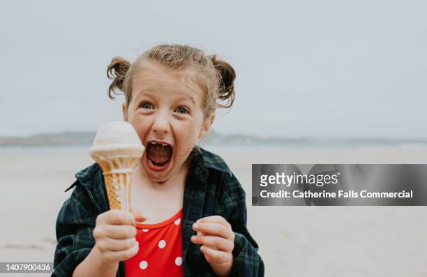 ecstatic little girl holds a vanilla ice-cream on a beach, looking delighted as she holds the cone towards the camera. - excited funny stock-fotos und bilder