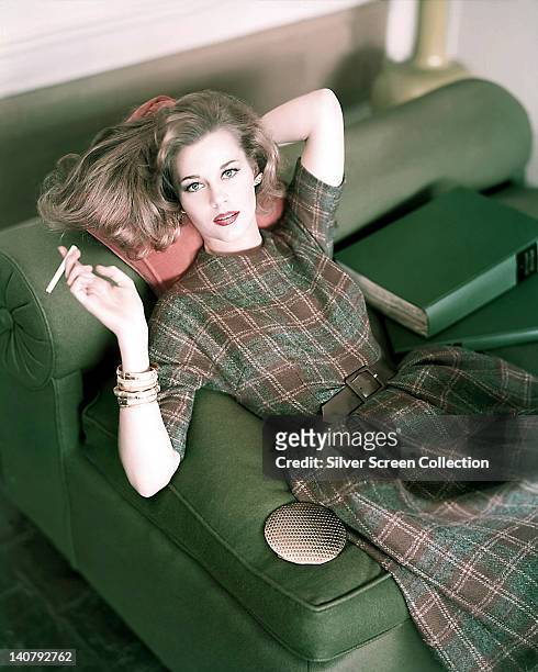 Jane Fonda, US actress, wearing a green and brown large check dress, posing with a cigarette and reclining on a green sofa, resting her head on a...