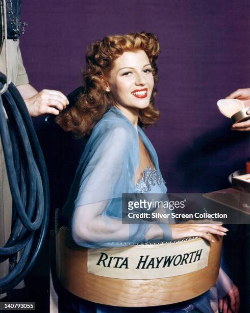 Rita Hayworth , US actress and dancer, wearing a sheer light blue gown while sitting in a chair, with her name written across the back rest, in a...