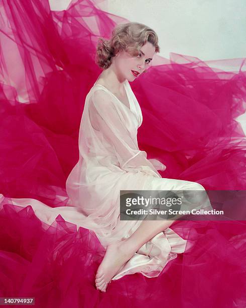 Grace Kelly , US actress, wearing a cream silk gown with sheer fabric arms, sitting on pink fabric in a studio portrait, circa 1955.