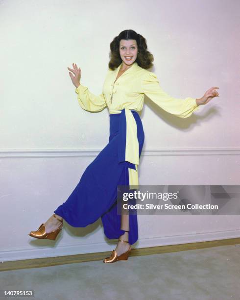 Rita Hayworth , US actress and dancer, wearing a yellow blouse and blue harem pants, holding a dance pose in a studio portrait, early to mid 1940s.