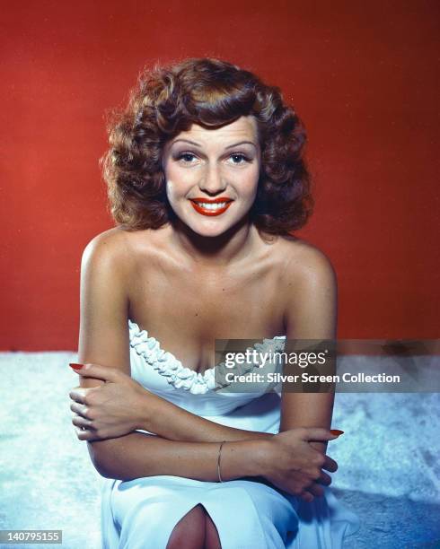 Rita Hayworth , US actress and dancer, wearing a white shoulderless dress and smiling in a studio portrait, against a red background, circa 1950.