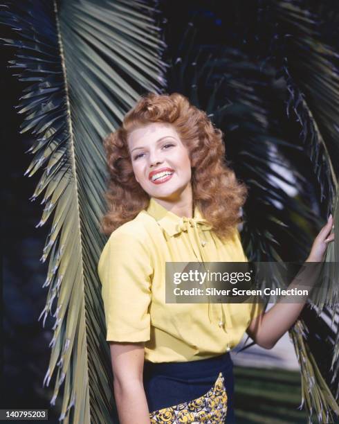 Rita Hayworth , US actress and dancer, wearing a short-sleeve yellow blouse, posing among palm fronds, circa 1945.