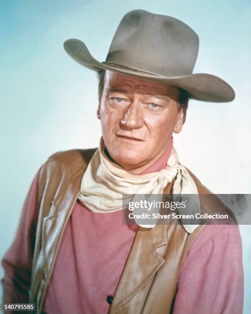 John Wayne , US actor wearing a tan leather waistcoat, a pink shirt and a white neckerchief, in a studio portrait, against a light blue background,...