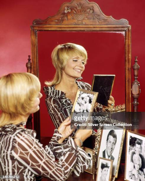 Florence Henderson, US actress, admiring photographs in frames on a table with a large mirror which reflects Henderson in a portrait issued as...