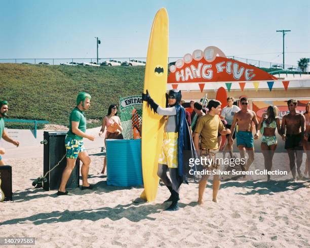 Adam West, US actor, wearing a pair of yellow and white long shorts over his costume, and standing on a beach with a yellow surfboard, with the...