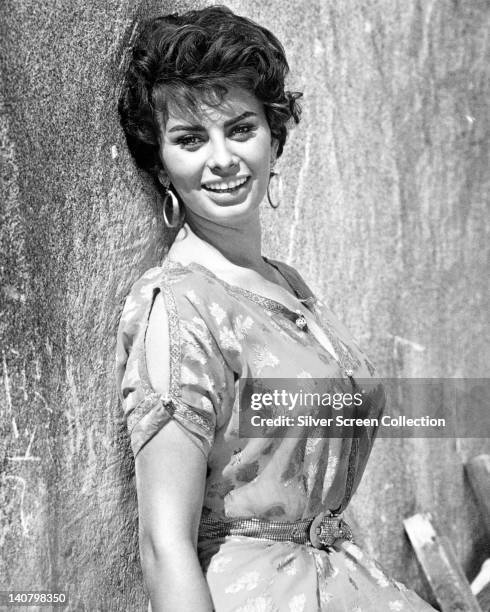 Sophia Loren, Italian actress, smiling and posing against a wall, wearing a short-sleeve print pattern dress, and large hoop earrings, circa 1955.