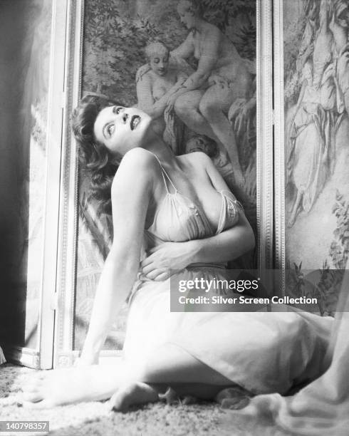 Tina Louise, US actress and singer, wearing a white thin strap gown, with her head thrown back, posing crouched on the ground against a printed...