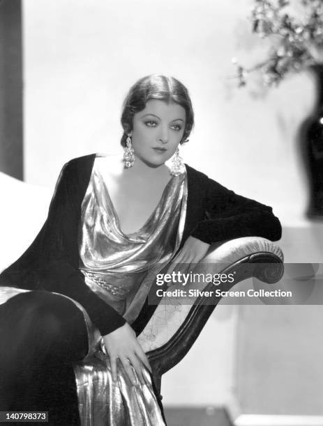 Myrna Loy , US actress, wearing a black cardigan over a silver dress, and large drop earrings, sitting with her arm resting on the arm of a sofa, in...