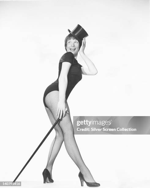 Shirley MacLaine, US actress, wearing a black leotard with fishnet stockings and a black top hat, posing with a cane in a studio portrait, against a...