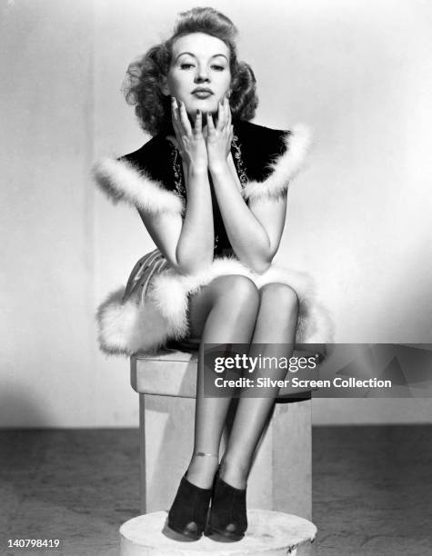 Jeanette MacDonald , US singer and actress, sitting on a pedestal, wearing a white fur-trimmed dress, with her chin resting in both her hands, circa...