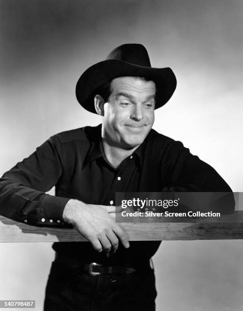 Fred MacMurray , US actor, with a moustache, leaning on a fence, and wearing a black shirt with a black cowboy hat, circa 1950.
