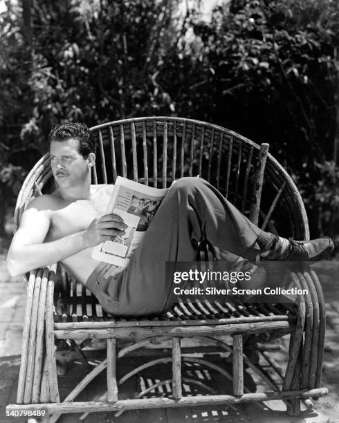 Fred MacMurray , US actor, with a moustache, reclining bare-chested in a wicker pool chair, reading a magazine, circa 1940.