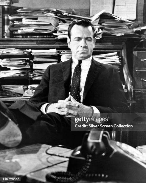Walter Matthau , wearing a black suit with his feet on the desk before him, with stacks of documents on the shelves behind him in an image issued...
