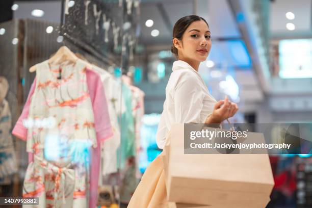 young asian woman holding shopping bag and looking at shop window while walking in shopping mall - shopping mall foto e immagini stock