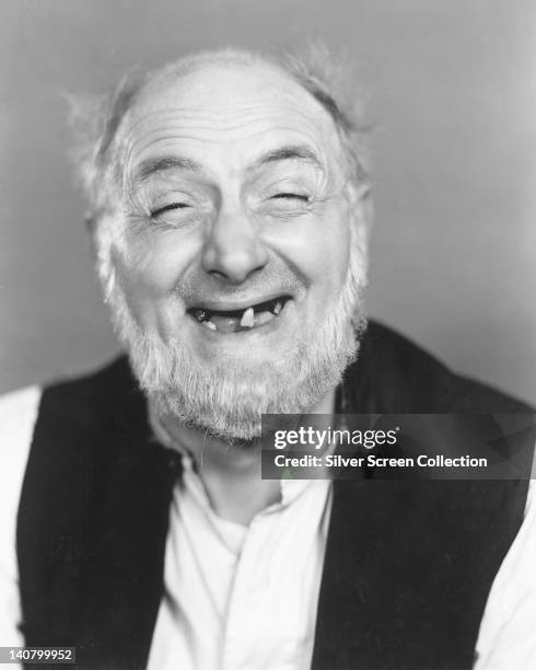 Moore Marriott , British actor, smiling with a toothless grin, wearing a black waistcoat over a white shirt in a studio portrait, circa 1940.