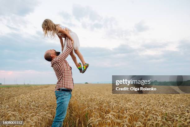 cheerful man, farmer in plaid shirt lifting little blonde girl up over the head in the air in the wheat field - horizon over land stock-fotos und bilder