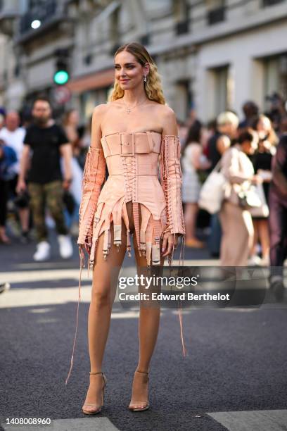 Chiara Ferragni wears silver and diamonds pendant earrings, a gold with cross pendant necklace, a beige shoulder-off / laces / corset short dress...