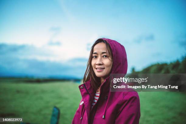 young asian woman in hoodie jacket smiling joyfully at the camera while relaxing in nature during a camping trip - un giorno nella vita foto e immagini stock